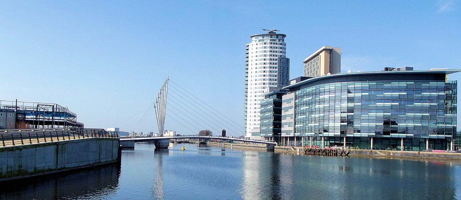 Photograph of a river in Salford, on a beautiful day, with large city buildings reflected in the water.